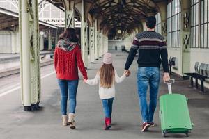 vista posteriore della famiglia amichevole tenersi per mano, portare la valigia, fare un viaggio, posare sul binario della stazione ferroviaria. giovane madre e padre, la loro piccola figlia aspettano l'allenamento e la partenza foto