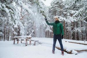 ritratto di sorridente ragazzo barbuto vestito con abiti caldi, scuote l'albero coperto di neve, ha un'espressione soddisfatta, ama trascorrere il tempo libero o i fine settimana con l'aria fresca d'inverno. concetto di stagione foto