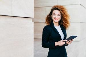 splendida donna in abito nero, con capelli ricci folti, occhi azzurri e labbra dipinte di rosso che sorride piacevolmente mentre digita un messaggio di testo sul tablet. imprenditrice di successo in abiti eleganti utilizzando tablet foto