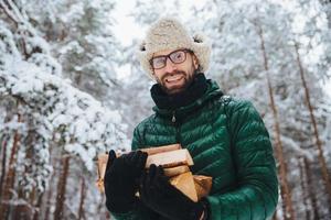 bel maschio barbuto con occhiali e cappello caldo con giacca a vento, tiene legna da ardere, posa contro alberi coperti di neve bianca scintillante, trascorre del tempo nella foresta. colpo esterno di uomo felice riposa nella foresta foto