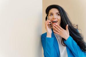 positiva donna d'affari apre la bocca per l'interesse, tocca il mento, guarda da parte, affascinata dalle voci, ha una conversazione telefonica con un caro amico, spettegola sulle ultime notizie, indossa un costume blu formale foto