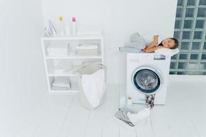 inquadratura di un bambino stanco dorme sulla lavatrice, usa un morbido asciugamano bianco come cuscino, fa sogni piacevoli, cestino e bacinella sul pavimento bianco, si sente affaticato dopo aver caricato i vestiti in lavatrice, essendo a casa foto
