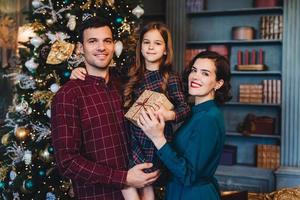 la famiglia felice sta insieme contro l'albero di abete decorato, guarda felicemente nella fotocamera, tieni presente, festeggia il nuovo anno nella cerchia familiare. bambina felice di trascorrere le vacanze con sua madre e suo padre foto