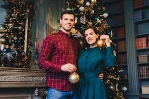 bell'uomo con stoppia abbraccia la sua bella moglie, tiene in mano palline di vetro, decora insieme l'albero di Capodanno, si prepara per le vacanze. sorridente giovane bella femmina e suo marito a casa foto