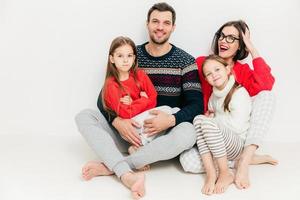 ritratto di famiglia felice di quattro membri attraente donna bruna, suo marito e due piccole figlie siedono sul pavimento, isolato su sfondo bianco. concetto di genitorialità. bella famiglia al coperto. foto