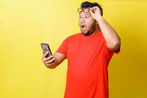 l'uomo grasso asiatico in maglietta rossa usa gli occhiali è scioccato e sorpreso quando guarda lo smartphone isolato su sfondo giallo foto