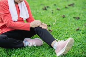 donna asiatica anziana con dolore alla caviglia del ginocchio mentre corre nel parco. donna asiatica senior che si siede per terra e che tiene il ginocchio doloroso. foto