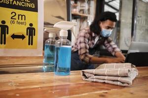 disinfettante per alcol per una protezione sana in un bar, barista asiatico maschio in maschera usa il laptop, in attesa di un ordine di caffè nella quarantena pandemica covid19, l'impatto economico per l'avvio di piccole imprese. foto