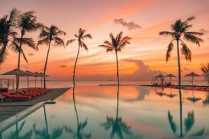 perfetto tramonto sulla spiaggia, piscina relax in un lussuoso hotel resort sulla spiaggia alla luce del tramonto banner perfetto per le vacanze al mare. paesaggio della spiaggia al tramonto. lettini palme riflessioni piscina a sfioro foto