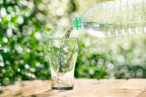 versare l'acqua da una bottiglia di plastica in un bicchiere. lo sfondo delle piante in giardino. focalizzazione morbida. foto