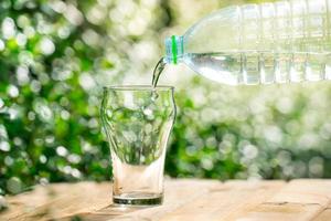 versare l'acqua da una bottiglia di plastica in un bicchiere. lo sfondo delle piante in giardino. focalizzazione morbida. foto