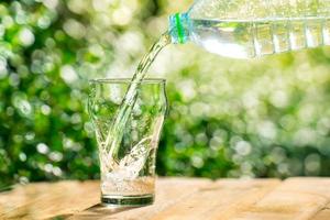 versare l'acqua da una bottiglia di plastica in un bicchiere. lo sfondo delle piante in giardino. focalizzazione morbida. foto
