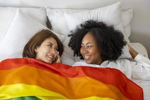 coppia dello stesso sesso matrimonio da razze diverse con bandiera arcobaleno lgbtq per il mese dell'orgoglio mentre si sdraiano insieme sul letto per promuovere l'uguaglianza e le differenze del concetto omosessuale foto