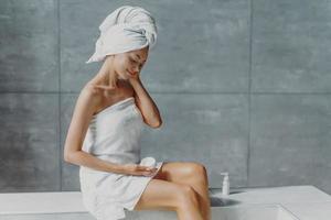 la giovane donna europea rinfrescata applica una crema antirughe, posa in bagno, avvolta in asciugamani da bagno, previene i segni dell'invecchiamento cutaneo, ha un corpo pulito dopo la doccia. benessere, concetto di benessere foto