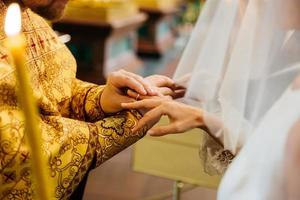 vista del sacerdote che indossa l'anello sulla mano della sposa, posa nella chiesa cristiana durante la cerimonia di matrimonio, candela accesa in primo piano foto