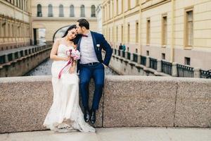 l'affettuosa coppia sposata si siede sul ponte insieme, si gode l'aria fresca, ha un buon rapporto, si bacia l'un l'altro. giovane donna in abito da sposa bianco riceve il bacio dal marito. concetto di stare insieme foto