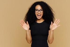 gioiosa giocosa donna afroamericana alza i palmi delle mani, si sente eccitata, esprime felicità, felice di ricevere regali, indossa abiti casual, occhiali, sta in piedi su sfondo marrone. reazione positiva foto