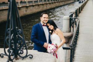 le coppie affettuose innamorate celebrano il loro matrimonio, posano davanti alla telecamera come stand vicino al ponte e al fiume, hanno sorrisi felici. felice coppia sposata positiva hanno relazioni romantiche. concetto di matrimonio foto