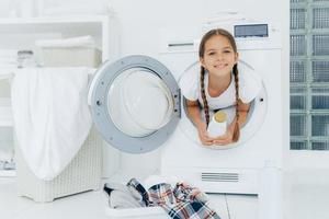 bambina sorridente con espressione felice, posa in lavatrice, vestita con una maglietta bianca, tiene in mano il detersivo, guarda con gioia la telecamera, circondata da molti vestiti e biancheria, fa i lavori domestici foto