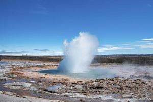 vista panoramica del geyser Strokkur che erutta in mezzo al paesaggio contro il cielo blu foto