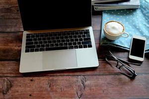 laptop con occhiali, smartphone, una tazza di caffè caldo e una rivista su un tavolo di legno, lavorando in remoto. foto