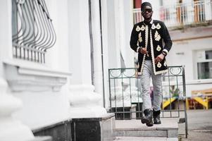 mega uomo africano alla moda nella tradizionale posa della giacca. ragazzo nero alla moda con cappello e occhiali da sole con il sigaro in mano. foto