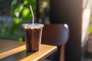 bicchiere di caffè freddo con latte, sciroppo di cioccolato in caffetteria. caffè freddo moka in tazza di plastica sul tavolo di legno in caffè con pianta e luce solare sullo sfondo foto