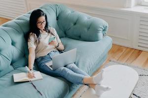 persone, apprendimento a distanza, tecnologie, concetto di lavoro freelance. una donna bruna seria scrive informazioni nel diario, tiene il telefono cellulare, si concentra sul lavoro a distanza, lavora con un computer portatile foto