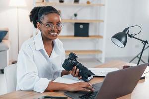 tenendo la fotocamera. la giovane donna afroamericana in camicia bianca è a casa foto