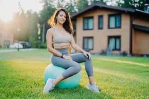 concetto di fitness, sport e allenamento. soddisfatta donna sportiva in buona forma fisica si prende una pausa dopo l'esercizio si siede sulla palla fitness all'aperto gode della natura e dell'aria fresca, fa esercizi di equilibrio pilates foto