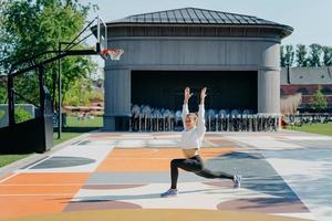 routine di allenamento personale. la donna atletica fa esercizi fisici all'aperto tiene le braccia alzate si riscalda prima di fare jogging vestita in pose attive sul campo da basket. flessibilità e forma fisica foto