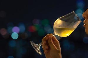 una persona che annusa il vino bianco in un bicchiere per la degustazione di vini con sfondo colorato di luci bokeh della città. foto