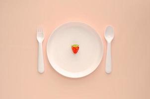 una fragola sul piatto bianco con forchetta e cucchiaio su sfondo rosa. concetto di cibo dieta sana creativa minima. foto