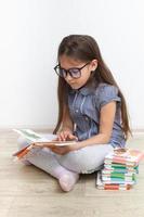 una bambina di 7 anni con gli occhiali legge un libro seduta per terra. educazione dei bambini, concetto di apprendimento foto