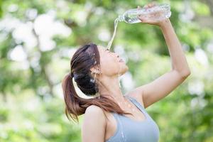 atleta giovane bella donna che beve e spruzza acqua in faccia al parco verde estivo, donna sportiva che beve acqua da una bottiglia di plastica dopo l'allenamento esercizio foto