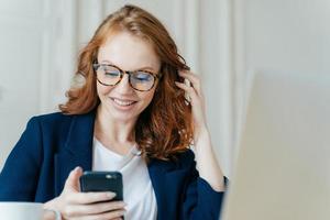 soddisfatta femmina dai capelli rossi utilizza un moderno dispositivo elettronico per controllare il feed di notizie, la comunicazione online e la ricerca di informazioni, si trova nello spazio di coworking, utilizza la connessione Internet ad alta velocità. foto