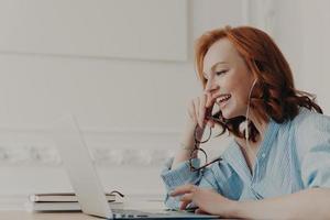 profilo di una donna rossa felice ride positivamente, comunica online con un amico, si prende una pausa dopo il lavoro, indossa una camicia blu, posa nello spazio di coworking. persone, lavoro autonomo, lavoro a distanza foto