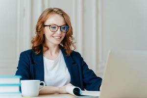 felice, la bella donna ha i capelli rossi, un sorriso positivo, si siede con un computer portatile sul desktop, è felice di fare videochiamate o conferenze, comunica con investitori dall'estero, beve caffè aromatico. foto