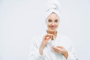 bella spa giovane donna con una pelle sana e fresca applica una lozione antietà o una crema cosmetica, usa una crema idratante da giorno, sta al coperto, vestita con accappatoio e asciugamano, fa la doccia prima di uscire foto