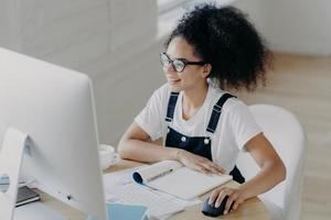 felice, la giovane donna afroamericana si siede davanti al computer moderno, fa il lavoro di progetto, ha molte carte e blocco note sul tavolo, indossa occhiali per la correzione della vista, vestita con abiti casual foto