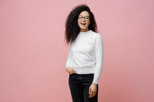 ripresa in interni di una donna piuttosto allegra che va al lavoro, ride di qualcosa di positivo, indossa un maglione bianco pulito e jeans, tiene la mano in tasca, si diverte con gli amici durante il tempo libero, posa in interni foto