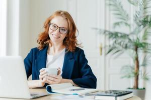 ripresa in interni di un editore femminile che lavora su un articolo, utilizza un computer portatile e Internet wireless, controlla le ultime notizie, tiene una tazza di caffè o tè, circondata da un blocco note per scrivere note, sorride felicemente foto