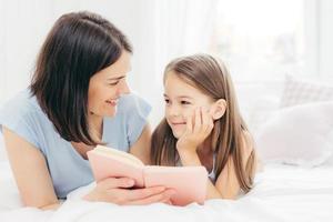 la piccola figlia contenta guarda incuriosita sua madre che legge fiabe, tiene in mano un piccolo libro, si sdraia su un letto comodo, si diverte insieme. il ragazzino allegro si prepara per la scuola. concetto di genitorialità foto