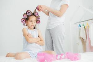 cupa bambina tiene le braccia conserte, ha i bigodini in testa, sua madre sta vicino e si fa i capelli, avvolge i bigodini, si prepara per la festa di compleanno, si prende cura della bellezza e dell'aspetto. concetto di infanzia. foto