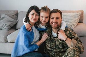 la bandiera ucraina è sulle spalle. il soldato in uniforme è a casa con la moglie e la figlia foto