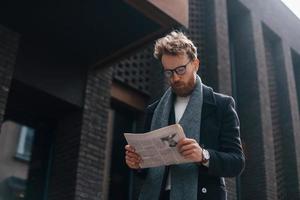 con il giornale in mano. l'uomo alla moda con la barba e con gli occhiali è all'aperto vicino all'edificio foto