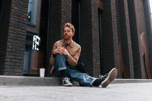 tazza di caffè. l'uomo alla moda con la barba in giacca color kaki e in jeans è all'aperto vicino all'edificio foto