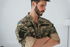 braccia incrociate. il soldato in uniforme è in piedi al chiuso contro il muro bianco foto