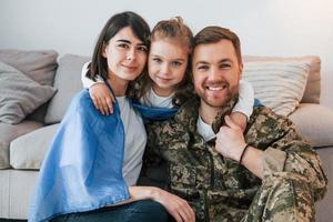 la bandiera ucraina è sulle spalle. il soldato in uniforme è a casa con la moglie e la figlia foto