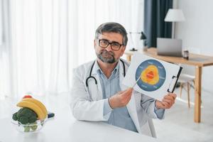 tenendo l'illustrazione del piano di dieta. l'operatore medico professionista in camice bianco è in ufficio foto